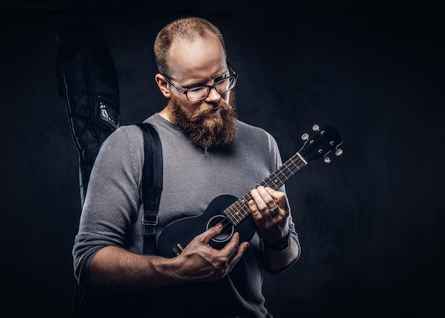 Redhead bearded male musician wearing glasses dressed in a gray t-shirt playing on a ukulele. Isolated on dark textured background.