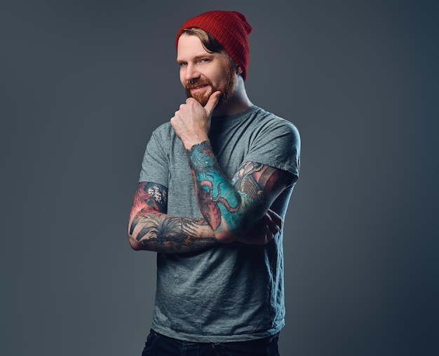 Redhead bearded male with tattoos on his arms posing over grey background.