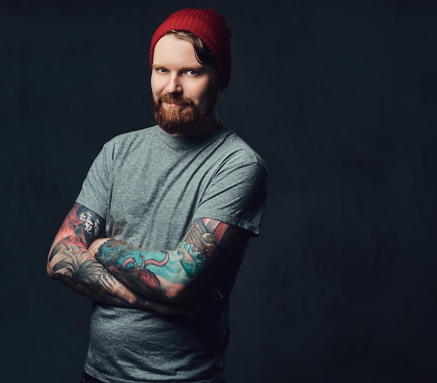 Redhead bearded male with tattoos on his arms posing over grey background.