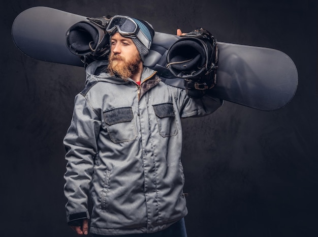 Free photo redhead bearded snowboarder in a winter hat and protective glasses dressed in a snowboarding coat posing with snowboard at a studio, looking away. isolated on a gray background.