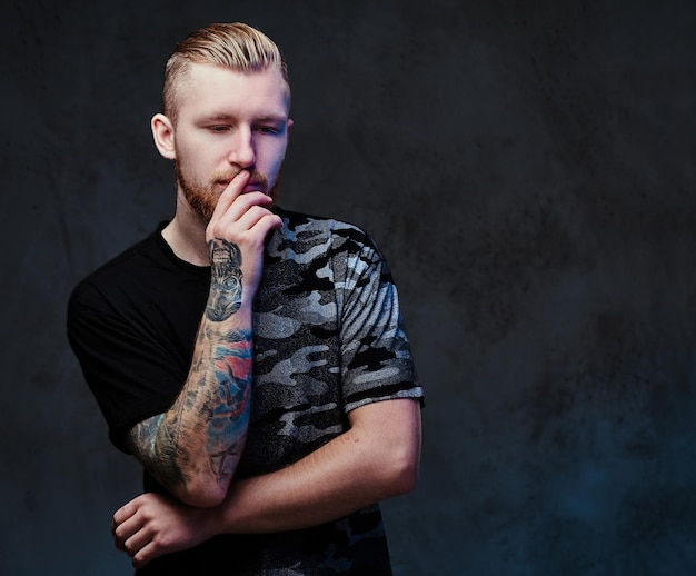 Free photo redhead bearded young male with tattoos on arms, dressed in a black t shirt over dark grey background.