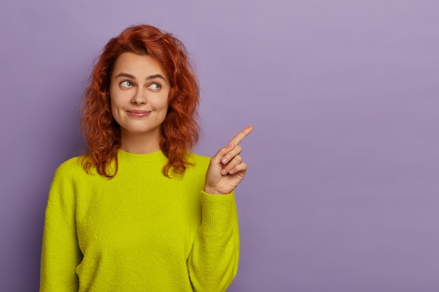 Free photo redhead beautiful woman points index finger aside, dressed in bright yellow jumper, demonstrates copy space for your advertising content or promotion, suggests using blank place for information