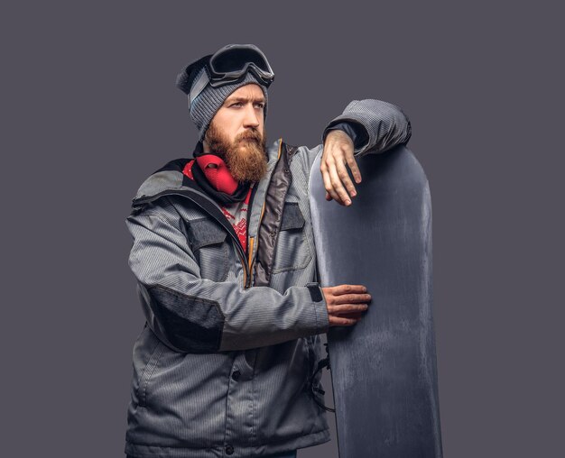 Redhead brutal snowboarder with a full beard in a winter hat and protective glasses dressed in a snowboarding coat posing with snowboard at a studio, looking away. Isolated on a gray background.