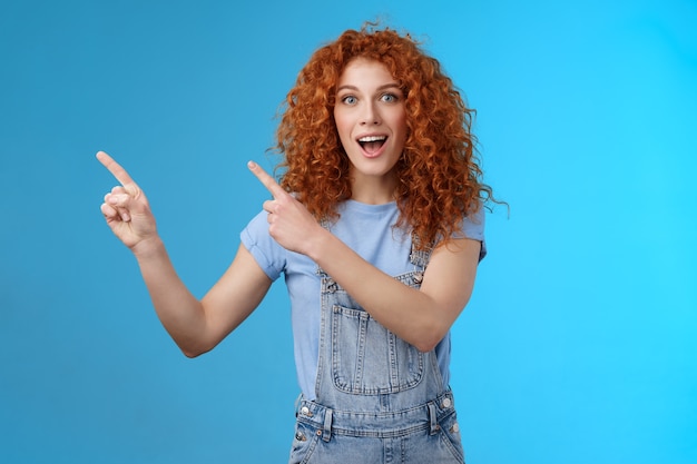 Redhead curly girl see awesome location take shot near beach social media vacation travel story pointing upper left corner amused look camera surprised enthusiastic smiling toothy excited grin.