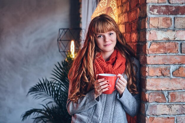 Redhead female in a red warm hat and gloves posing over the wall of a brick.