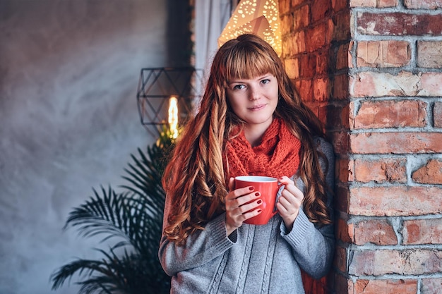 Redhead female in a red warm hat and gloves posing over the wall of a brick.