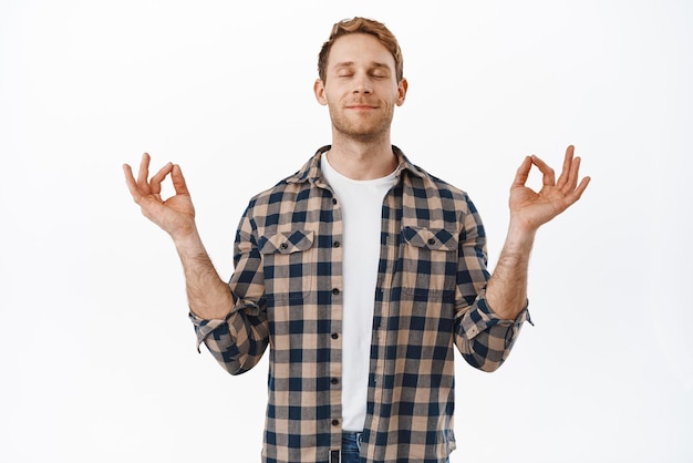 Redhead man close eyes and meditates with zen mudras smiling and breathing relaxed peaceful meditation or yoga trying to calm down standing over white background