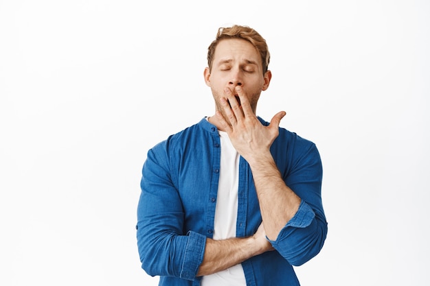 Free Photo redhead man feels tired and yawns, covers mouth with palm and close eyes, exhausted or bored, stands against white wall in casual clothes