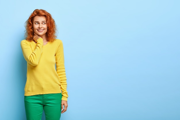 Free photo redhead positive woman has lucky upbeat mood, keeps hand on neck, has dremy happy look aside, wears yellow and green outfit