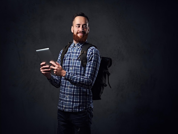 The redhead traveler hipster male holds tablet PC and a backpack.