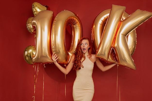 Free photo redhead woman in elegant dress posing excited holding golden balloons on red