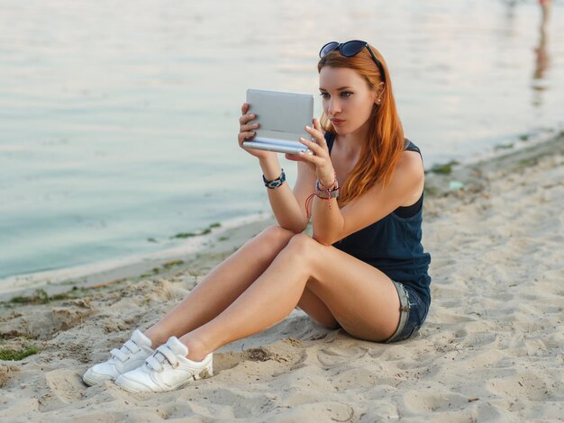 Redhead woman in jeans shorts and blue t shirt sitting on a beach and holding tablet computer.