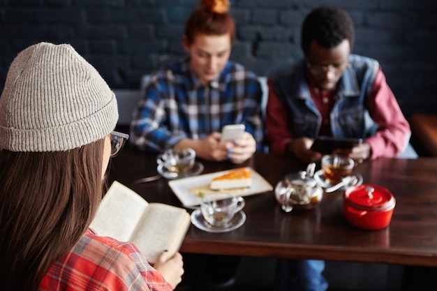 Free photo redhead woman making online order while shopping via internet on mobile telephone while having lunch in modern cafe interior with friends. selective focus on unrecognizable woman who is reading book