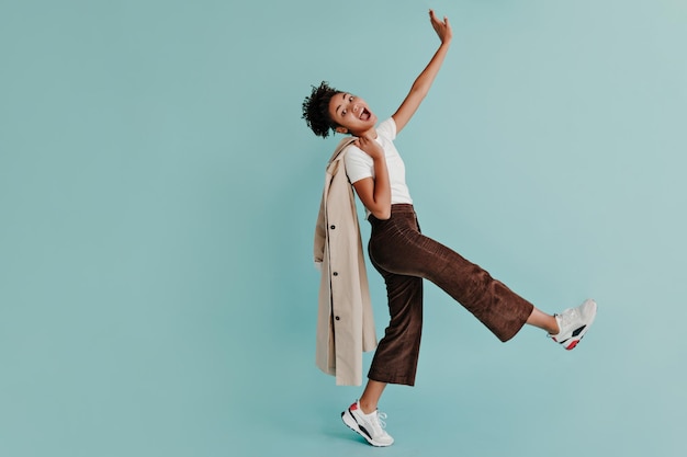 Refined african girl standing on one leg and waving hand on turquoise background Full length view of smiling black woman with trench coat