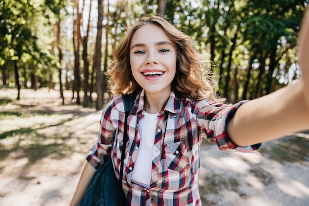 Free photo refined caucasian girl in checkered shirt walking in forest. outdoor portrait of laughing curly lady making selfie in sunny day.