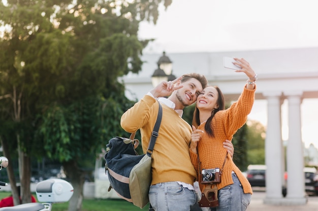 Refined dark-haired woman making selfie with handsome man in orange shirt with white achitecture on background