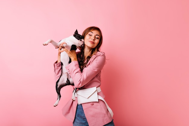 Refined girl smiling with eyes closed while posing with cute dog. Indoor portrait of ginger lady having fun with french bulldog.