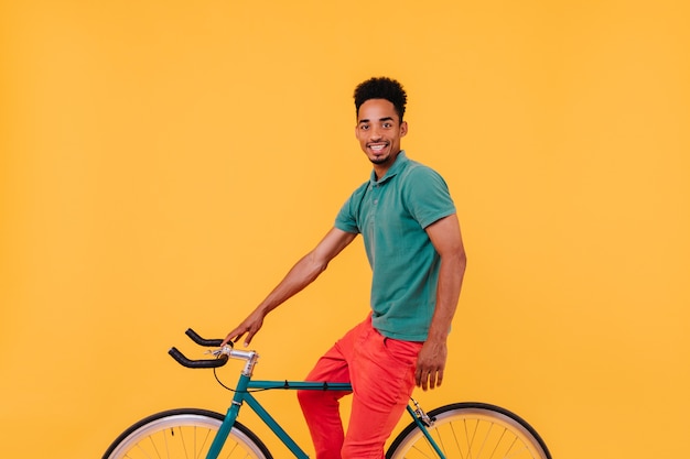 Refined male cyclist posing with smile. Interested brunette guy sitting on bicycle near yellow wall.