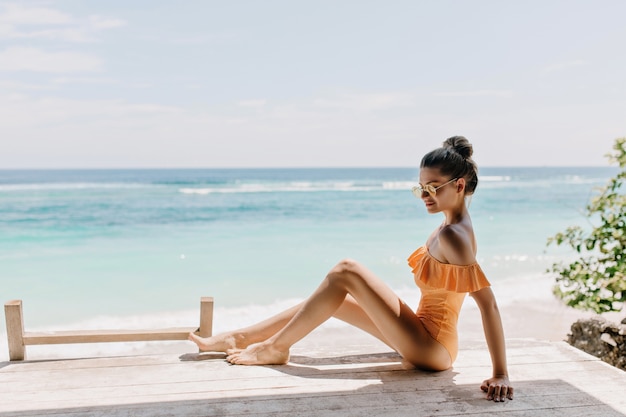 Refined slim young woman posing in the beach.  Pleasant tanned girl in sunglasses and stylish swimsuit chilling on ocean coast with shy smile.