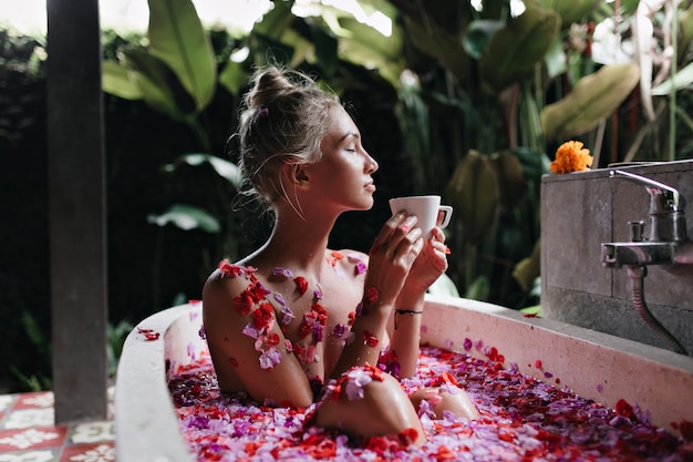 Refined woman sitting in bath on nature background. Wonderful caucasian lady relaxing during spa and drinking tea.