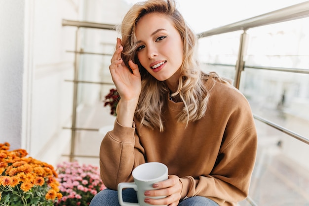 Refined young woman drinking tea at balcony. Pretty fair-haired girl enjoying coffee in morning.