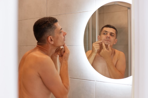 Free photo reflection in the mirror handsome dark haired male standing with naked upper body and looking at his face, finds pimple, having skin problems, morning hygiene procedures.