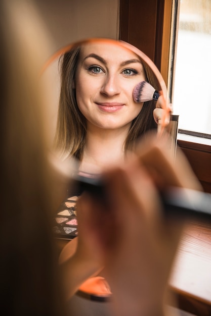 Free photo reflection of a smiling woman applying blusher on her face