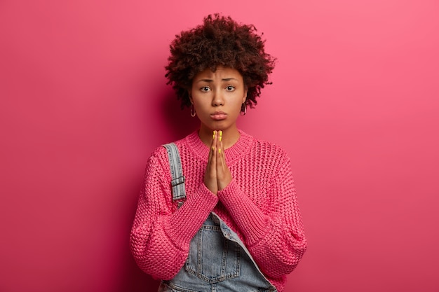 Regretful dark skinned African American woman beggs for forgiveness, feels guilty, purses lower lip, wears knitted sweater, isolated on pink wall, beseeches parents for money, looks innocent