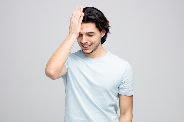 regretful young handsome man keeping hand on head looking down isolated on white background