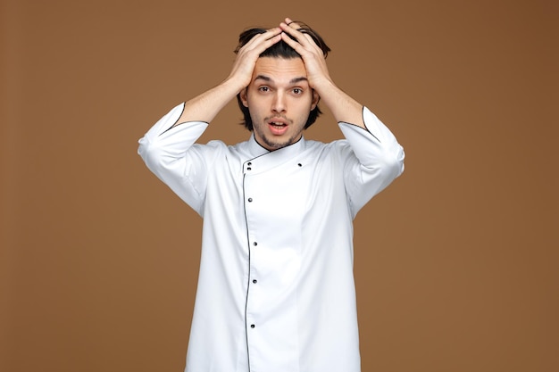 Free photo regretful young male chef wearing uniform keeping hands on head looking at camera isolated on brown background