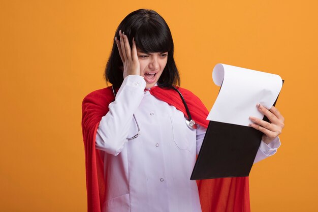 Regretted young superhero girl wearing stethoscope with medical robe and cloak flipping through clipboard putting hand on head isolated on orange wall