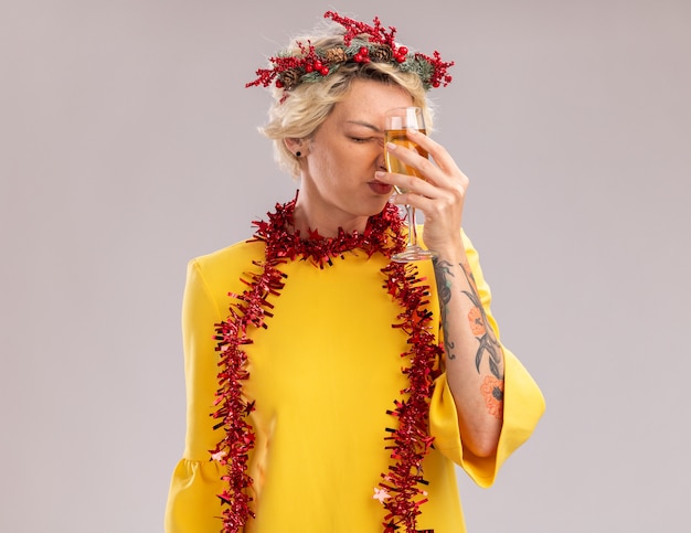 Free photo regretting young blonde woman wearing christmas head wreath and tinsel garland around neck touching forehead with glass of champagne with closed eyes isolated on white wall with copy space