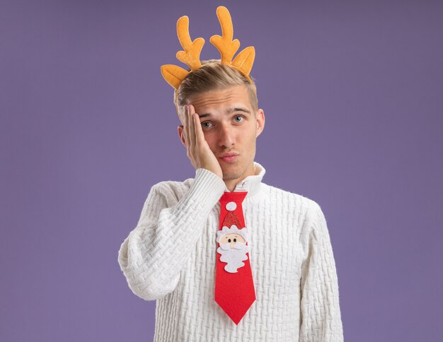 Regretting young handsome guy wearing reindeer antlers headband and santa claus tie looking at camera keeping hand on face isolated on purple background with copy space