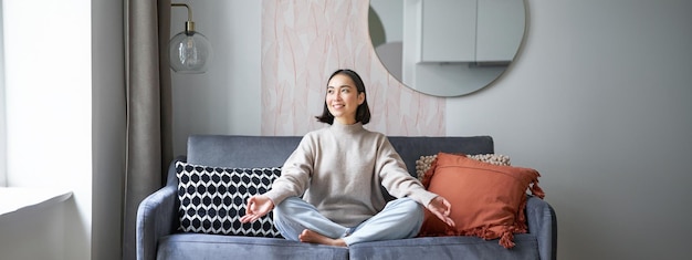 Relaxation and patience smiling young asian woman in cozy room sitting on sofa and meditating doing