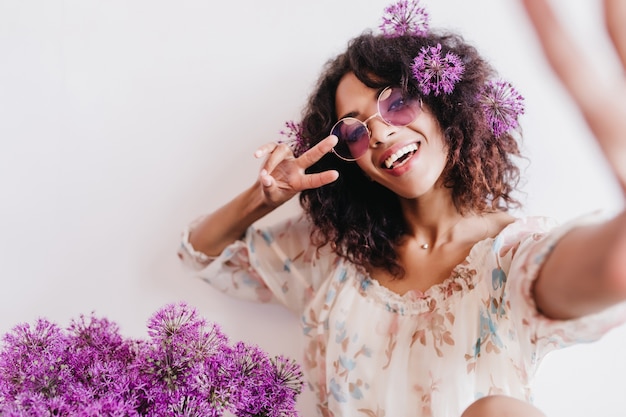 Relaxed african girl taking picture of herself with purple alliums. Indoor photo of fascinating curly female model in sunglasses.