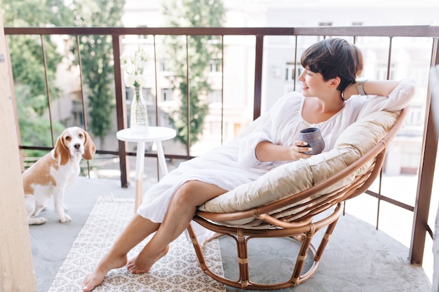 Free photo relaxed barefooted girl in white dress sitting in chair on balcony and holding cup of tea