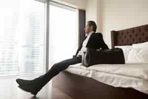 Free photo relaxed businessman sitting on bed besides luggage bag.