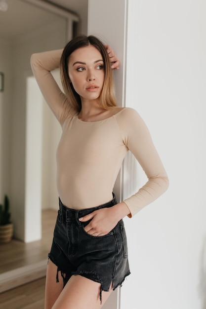 Relaxed cheerful blond woman in stylish beige pullover and shorts smiles sincerely and looking aside Happy pretty girl stands over white wall