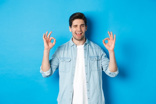 Free photo relaxed and confident man showing ok signs and winking, everything okay gesture, standing against blue background.