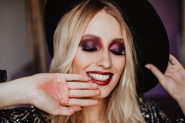 Free Photo relaxed female model with shiny makeup smiling with eyes closed. pleased gorgeous girl with long blonde hair posing in hat.