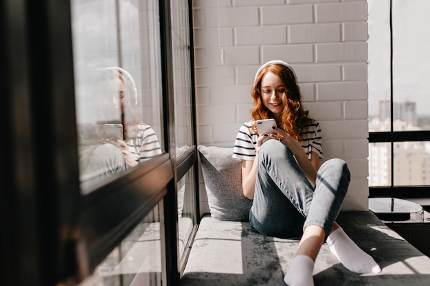 Free photo relaxed girl in jeans sitting on window sill. magnificent ginger lady in headphones holding phone.