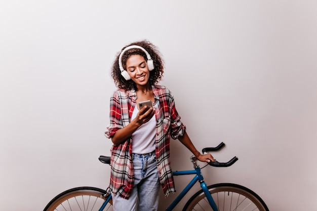 Relaxed girl with phone standing near bicycle and smiling. Charming african woman listening music and texting message.