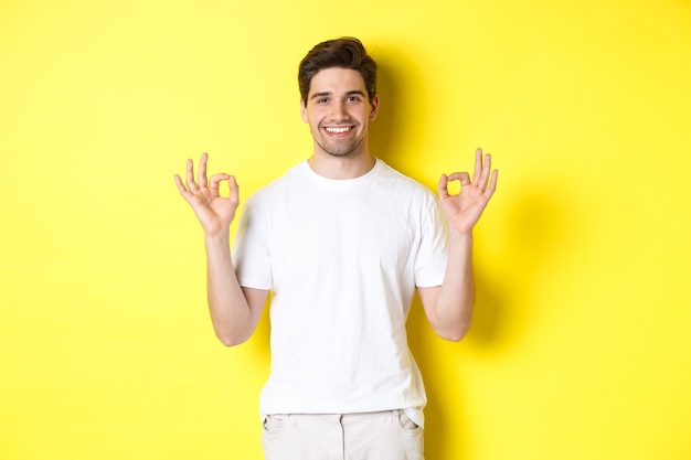 Relaxed guy smiling, showing okay signs, approve or agree, standing against yellow background