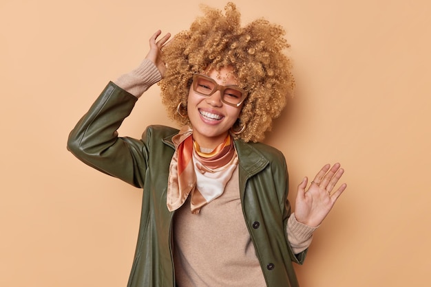 Free photo relaxed happy woman with curly bushy fair hair stands over beige background dances happily to music lifts arms dressed in fashionable clothes smiles broadly feels free and carefree enjoys life