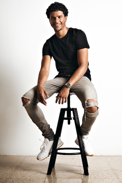 Free photo relaxed smiling athletic young model in torn jeans, white sneakers and plain black t-shirt on bar stool with white walls