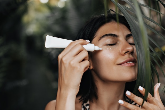 Relaxed woman applying sunscreen. Joyful woman with face cream standing on nature background.