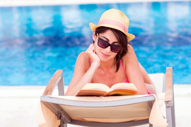 Relaxed woman reading a book with swimming pool background