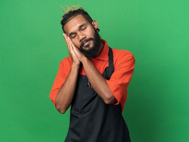 relaxed young afro-american male barber wearing uniform doing sleep gesture isolated on green wall with copy space