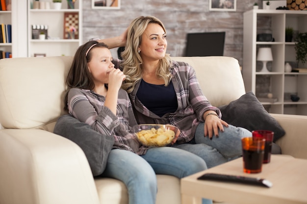 Relaxed young mother and cheerful daughter watching tv sitting on sofa eating chips.