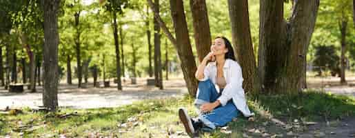 Free photo relaxed young woman resting near tree sitting in park on lawn under shade smiling and looking happy